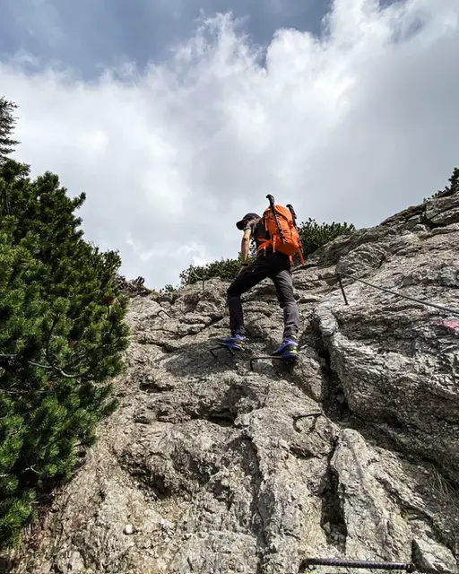 Der Tester trägt den Bergtagen 38 in orange und passiert gerade einen kurzen mit Eisenstangen und Stahlseil versicherten Wegabschnitt. Die Wanderstöcke sind am Rucksack befestigt.